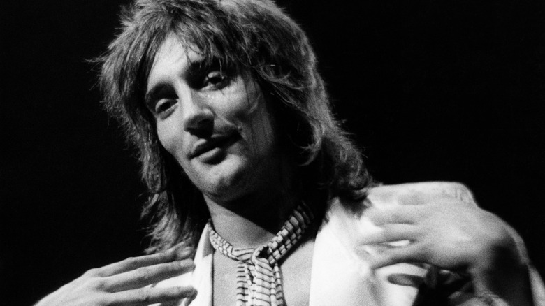 A black and white photo of Rod Stewart smiling while gesturing toward himself