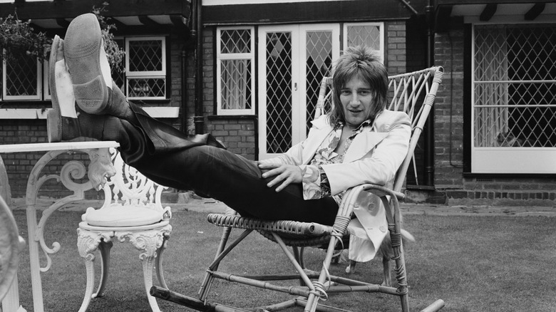 A black and white photo of Rod Stewart kicking his feet up on a table while sitting in a rocking chair