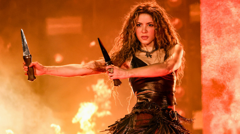 Shakira holding two swords with a fire blazing behind her on stage