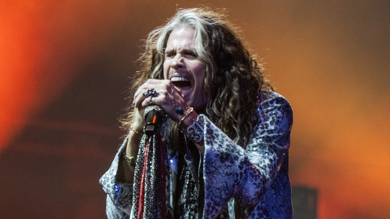 Steven Tyler screaming into a microphone with both hands during a 2023 performance