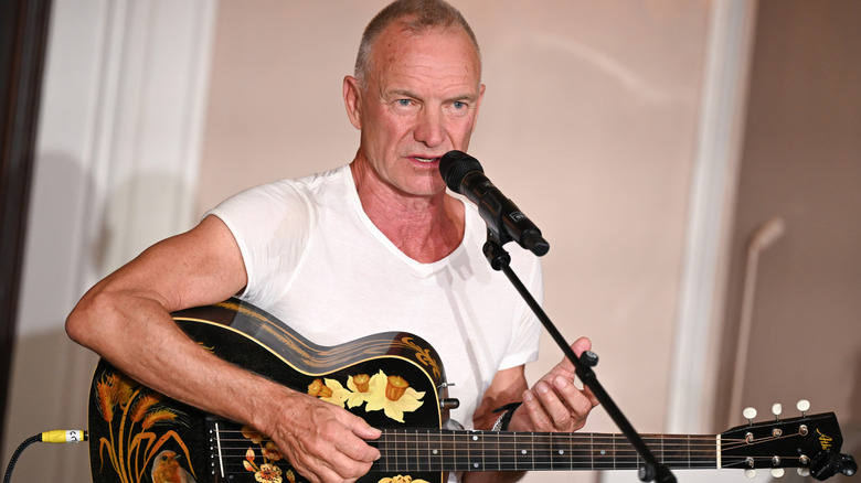 Sting sitting singing and playing the guitar in white shirt
