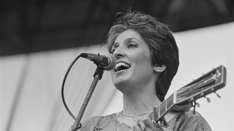 Joan Baez smiling onstage and singing in front of microphone while playing guitar
