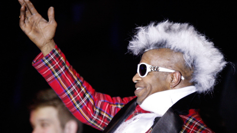Sly Stone wearing sunglasses and red tartan suit smiling with raised hand