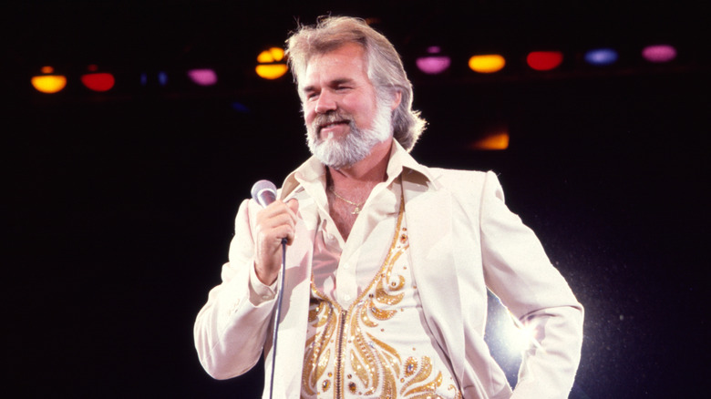 Kenny Rogers wearing a white and gold suit on stage, holding a microphone.