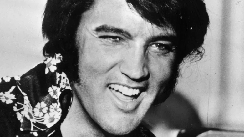 A black and white image of Elvis Presley. He is wearing a Hawaii shirt with flowers and has black and wavy hair and is
smiling.