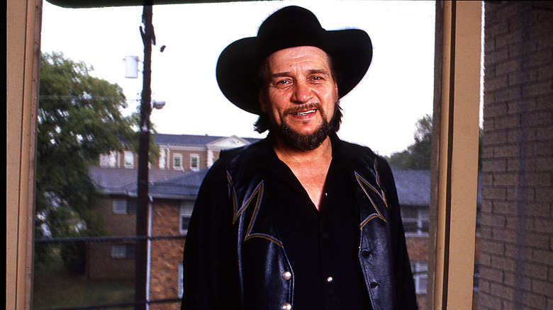 Waylon Jennings smiles in his office while wearing a black hat and black jacket