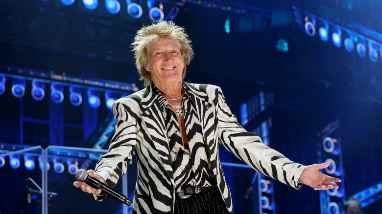 Rod Stewart performing on stage, with his arms wide open, in 2025