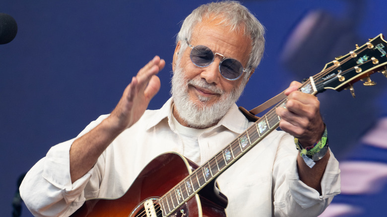 Cat Stevens wearing purple sunglasses and white shirt strumming a guitar on stage