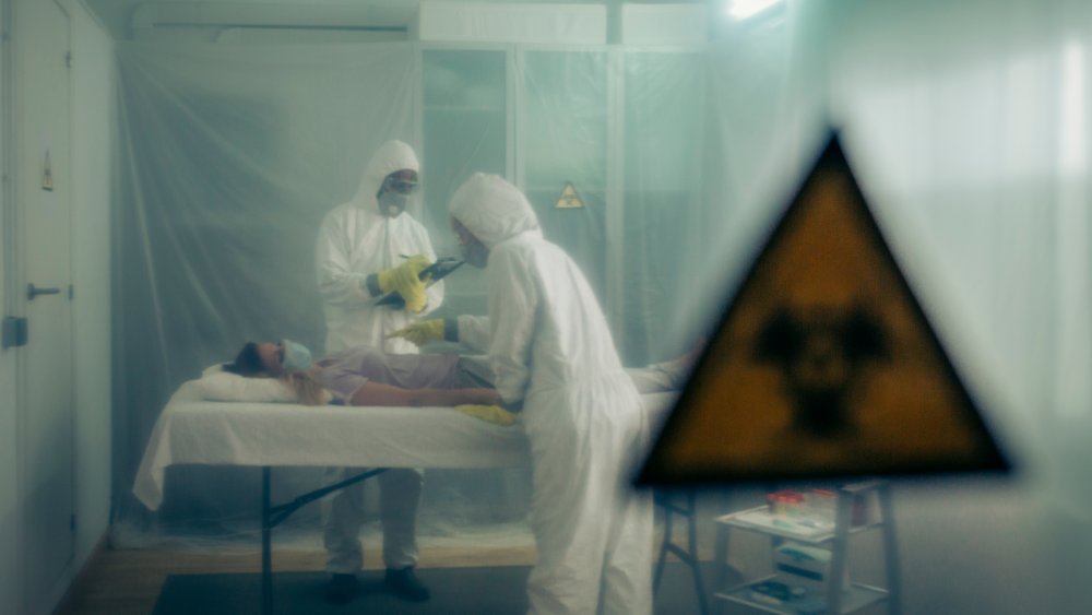 Two people attending to a woman with a virus lying on a stretcher in a field hospital with bio hazard symbol in the foreground