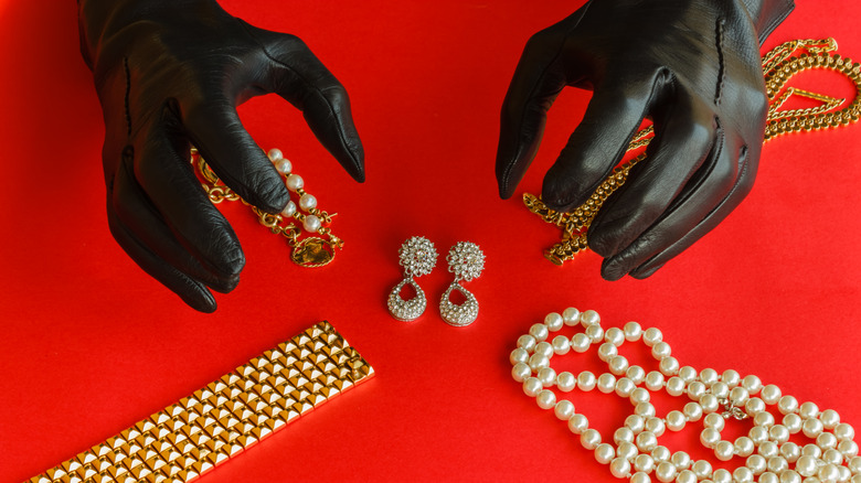 Two hands wrapped in black gloves reach towards jewlery against a red background.