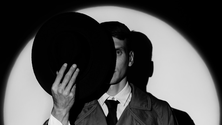 Black and white photo of man in vintage coat, face partially shielded by hat held in front.