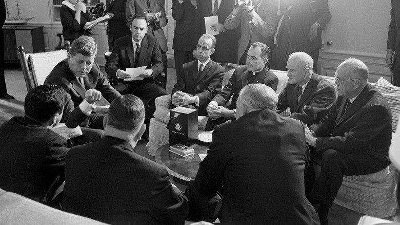 Kennedy seated with aides and legislators surrounded by press photographers