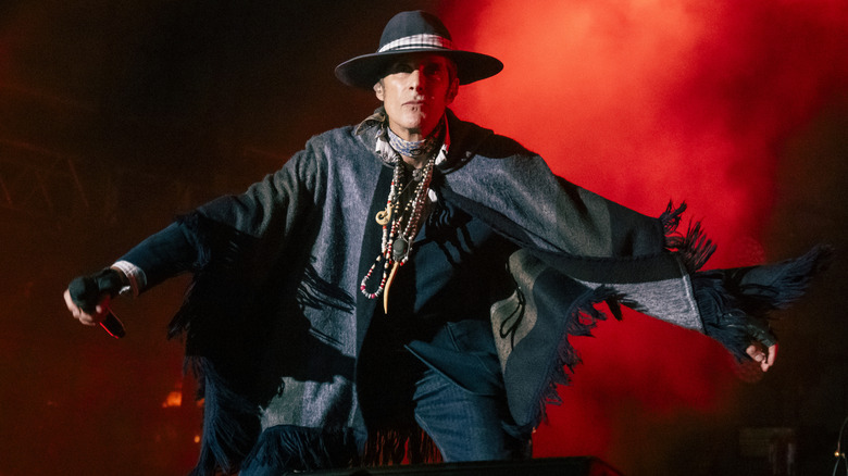 Perry Farrell in hat and poncho wearing beads on red-lit stage