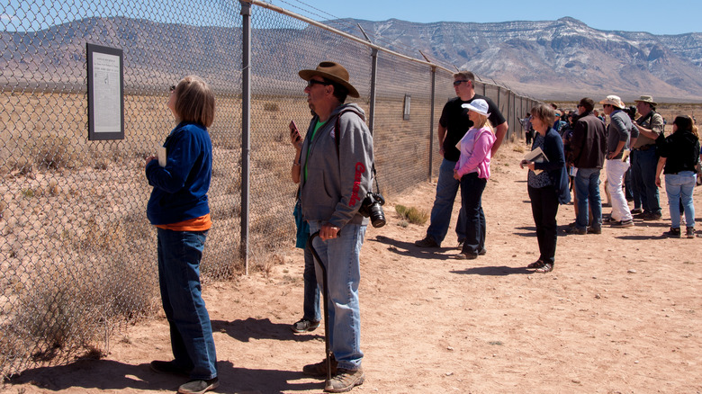visitors trinity test sit stand next to chain link fence