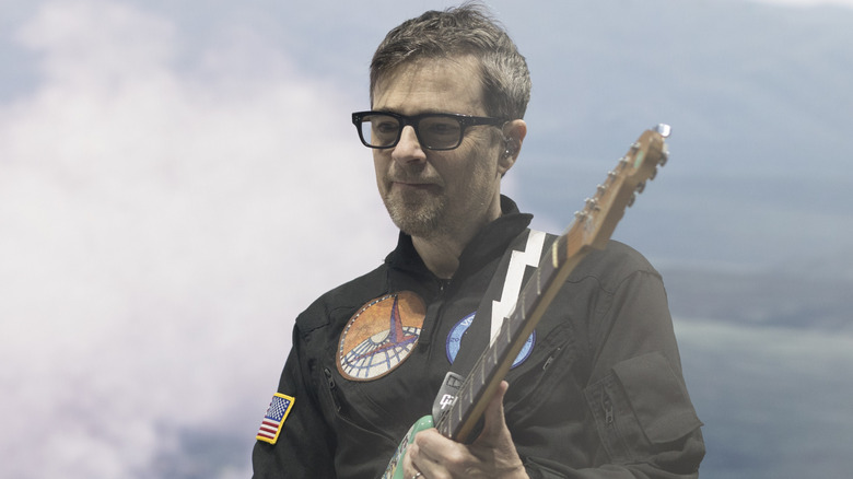 Rivers Cuomo looking off sadly while playing in front of clouds