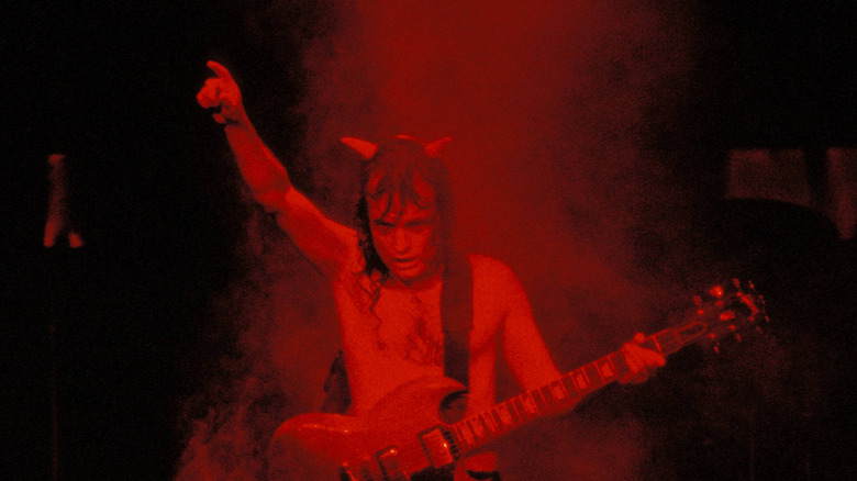 Angus Young wearing a devil horn headband under red stage lights while holding his guitar with one arm up