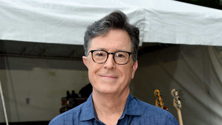 Stephen Colbert wearing a blue shirt, posing in front of a tent