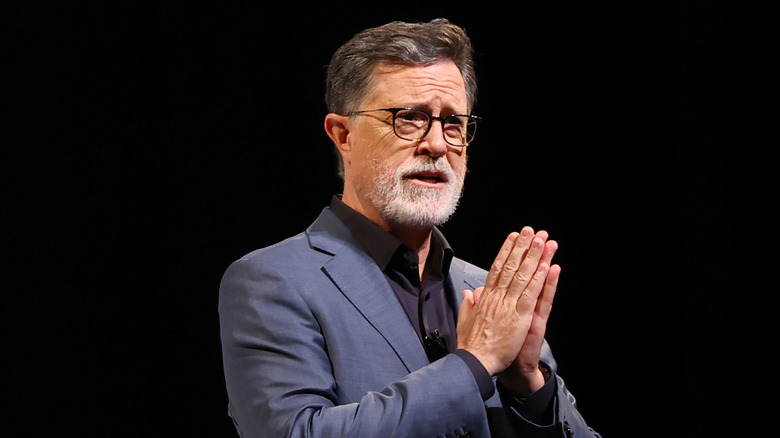 Stephen Colbert with a grey beard clasping the palms of his hands together in front of a black background