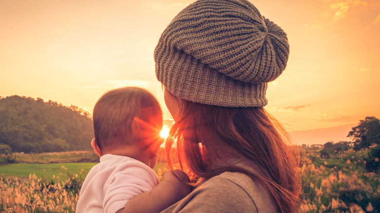 Young mother standing near a river & carrying her baby looking at the beautiful Sunset