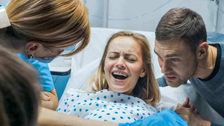 woman in labor screaming on hospital bed with man and medics