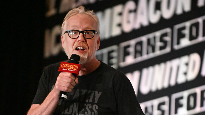 Adam Savage speaking in front of a wall of words