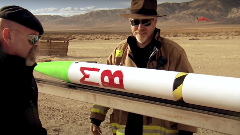 Adam Savage and Jamie Hyneman in hats and sunglasses standing in desert by a model rocket