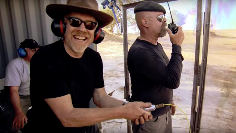Adam Savage with goatee and sunglasses smiling next to Jamie Hyneman at a range