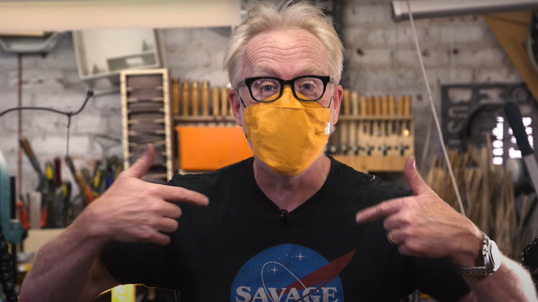 Adam Savage in glasses and yellow mask pointing to himself in workshop