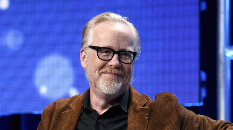 Adam Savage with goatee and glasses sat smiling in tan jacket