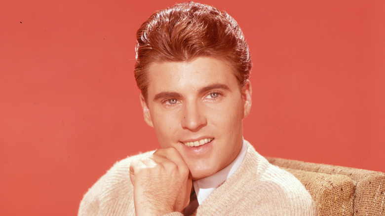 Ricky Nelson wearing a cream sweater and posing with his hand on his chin in front of a red backdrop in the 1950s.