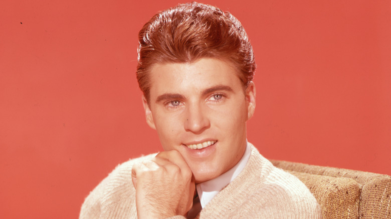 Ricky Nelson wearing a cream sweater and posing with his hand on his chin in front of a red backdrop in the 1950s.