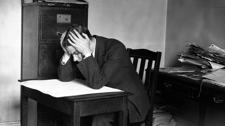 Man at desk stares at paper with head in hands, black and white image.