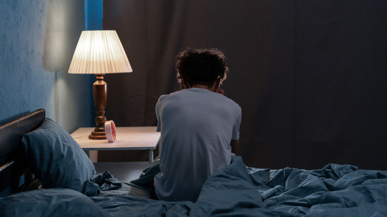 Person sitting on side of bed silhouetted by lamp, seen from behind.