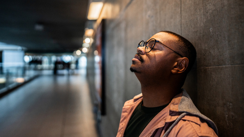 Man with glasses closes eyes and leans against wall.