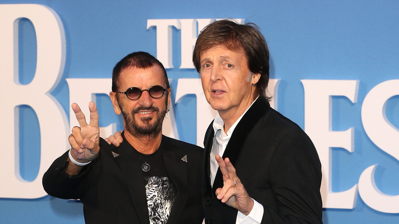Ringo Starr flashing the peace sign while standing next to Paul McCartney in front of the Beatles logo
