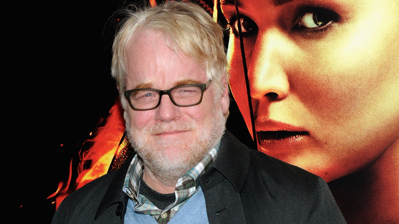 Philip Seymour Hoffman smirking in glasses in front of Hunger Games poster