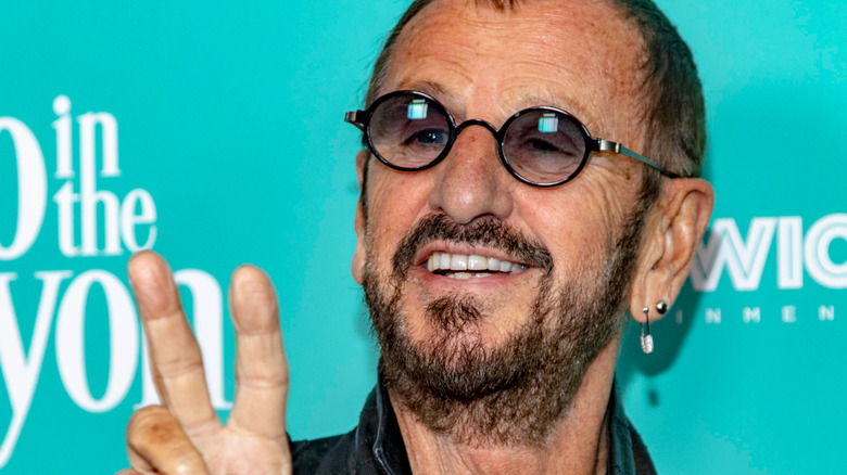 Ringo Starr smiling in glasses making peace sign blue background