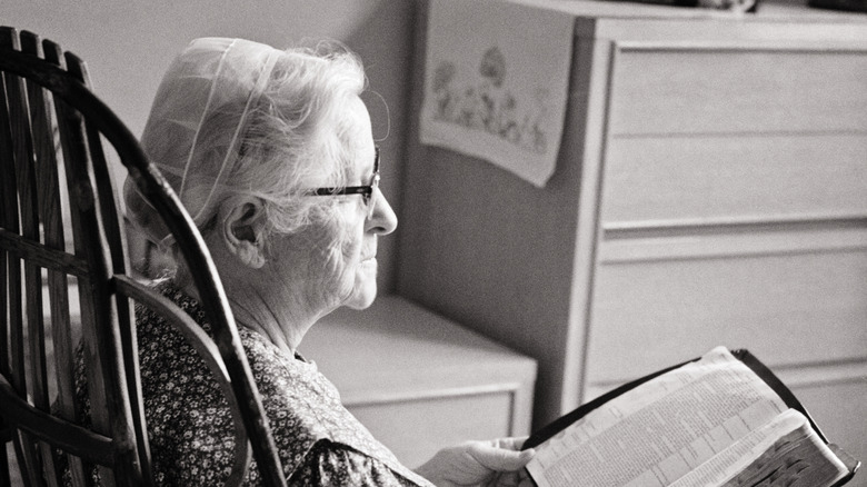 An elderly Amish woman sitting in a chair and reading the Bible.
